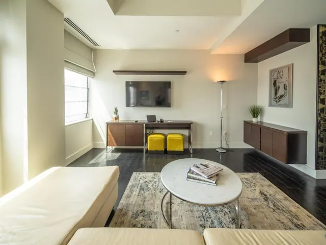 A living area with a cream sofa, a round marble coffee table holding books, two yellow ottomans, a wall-mounted television, a floor lamp and a wood sideboard.
