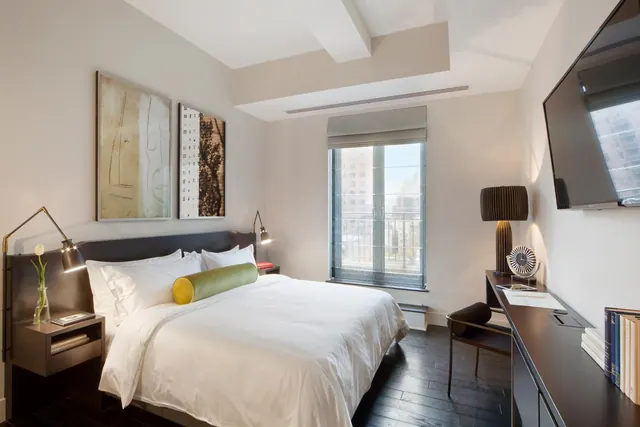 A bedroom with a king bed dressed in white linens, two framed artworks, wall lamps, a window onto nearby city buildings, and a wall-mounted television beside a desk.