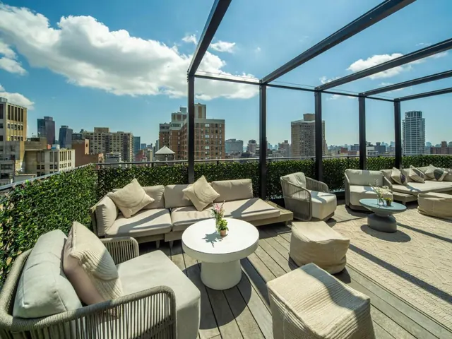 A rooftop terrace with outdoor sofas, armchairs and low tables under a metal pergola, with city buildings visible beyond the hedge wall.