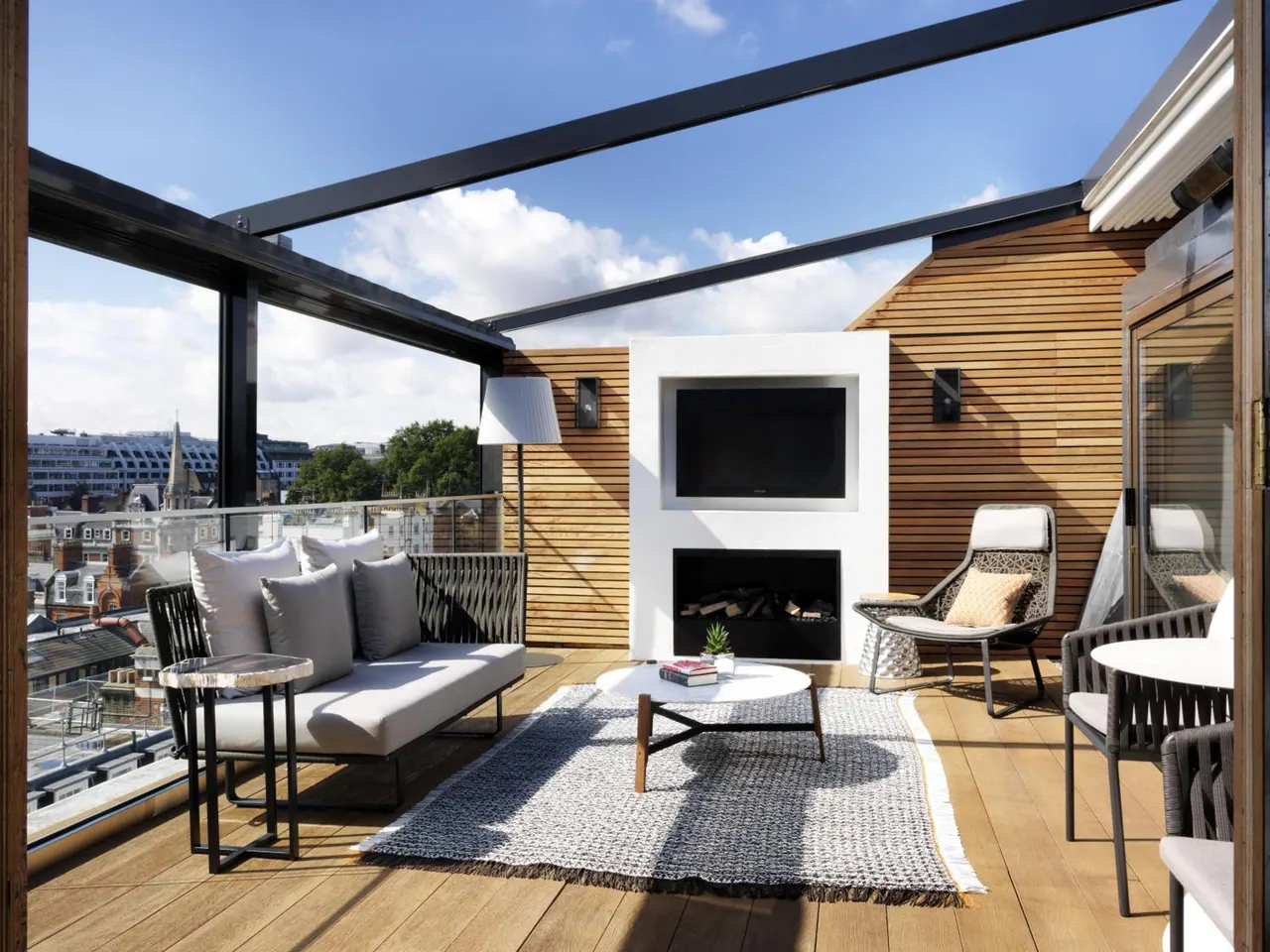 A covered rooftop terrace with a grey sofa, armchairs, a wall-mounted television above a fireplace, and a view over surrounding rooftops.