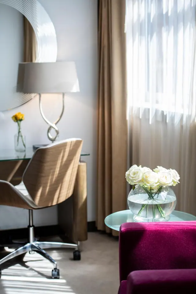 A hotel room corner shows a wooden desk chair, a desk lamp, a small vase of yellow flowers, a glass side table with white roses, and a magenta sofa beside sheer-curtained windows.