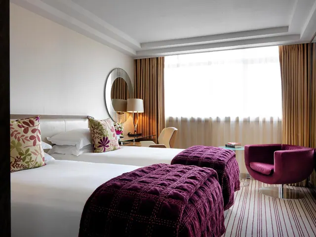 A hotel bedroom with two beds dressed in white linen and plum throws, a desk with a lamp and chair, and a plum armchair beside a window with sheer curtains.