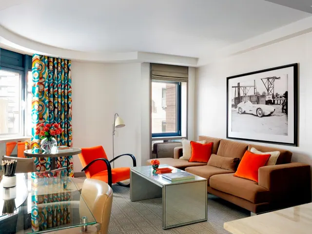 A living area with an orange armchair, a brown sofa with orange cushions, a glass-topped table and a framed black and white photograph on the wall.