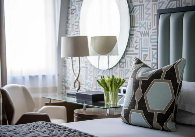 A bedroom with an upholstered teal headboard, a patterned cushion on the bed, a round mirror, a table lamp, a white armchair and a glass side table holding flowers.