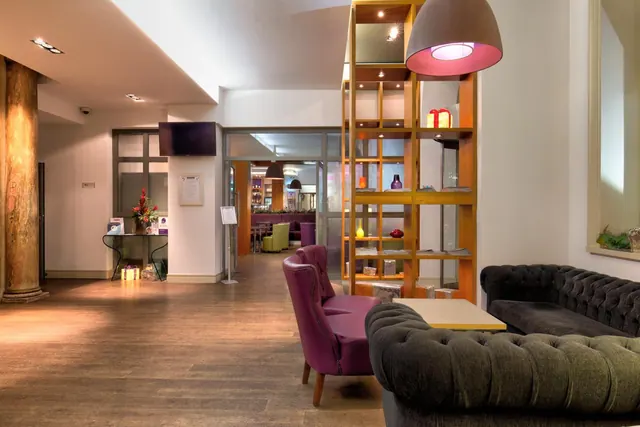 A lobby seating area with a dark tufted sofa, purple armchairs, a wooden shelving divider and a wall-mounted television.