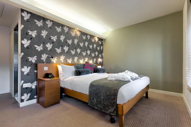 A double bed with white bedding and folded white robes sits beside a patterned grey feature wall and a wooden bedside table in a hotel bedroom.