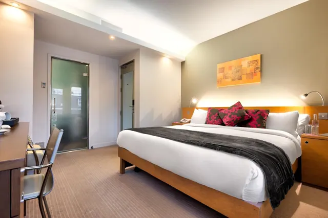 A double bed with a dark throw and red cushions sits beside a wooden bedside table, with a desk and chairs on the left in a hotel bedroom.