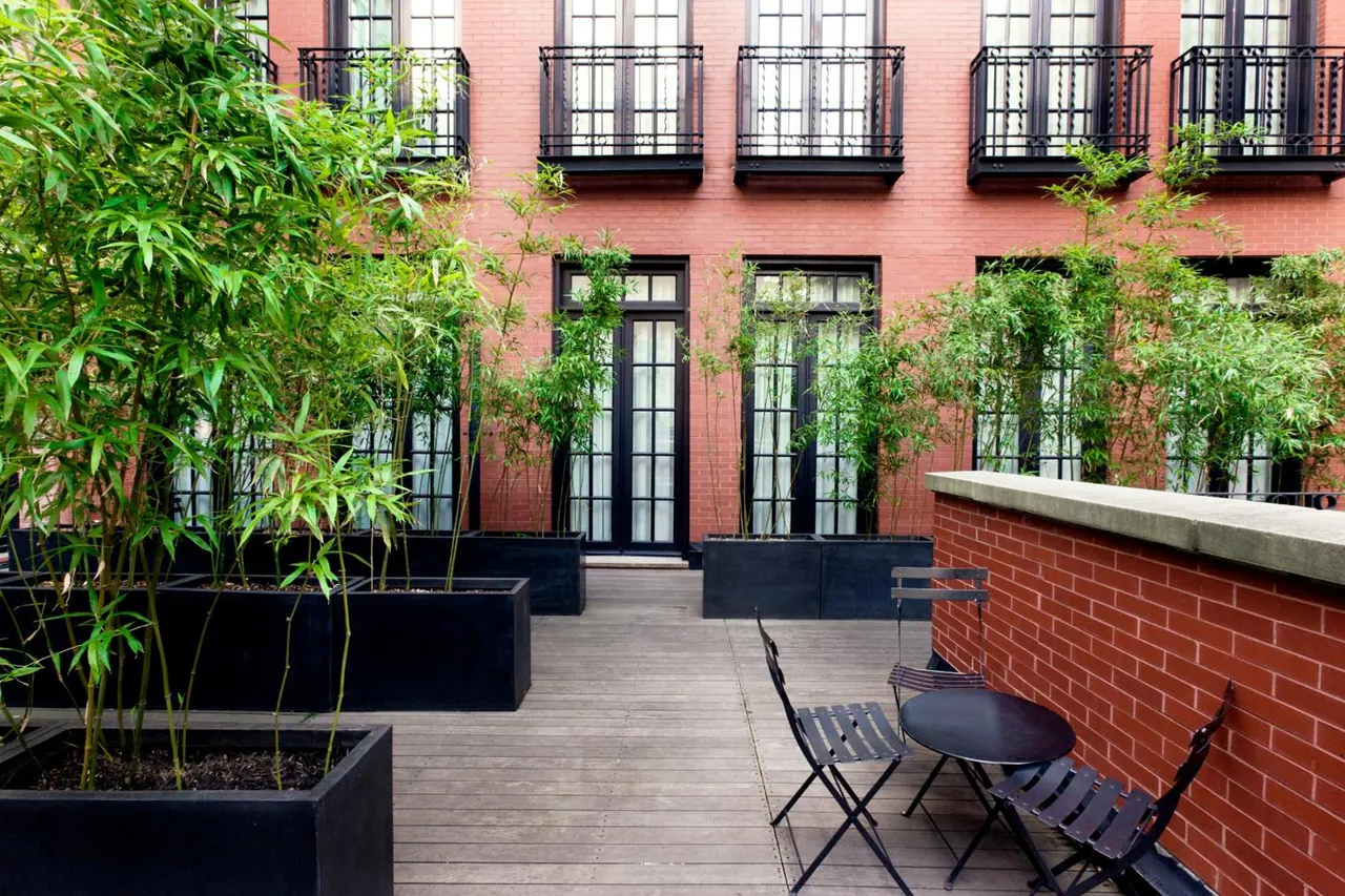 A brick-walled courtyard terrace with a wooden deck, black planters of bamboo, folding bistro chairs and a round table, with French doors and Juliet balconies on the building behind.