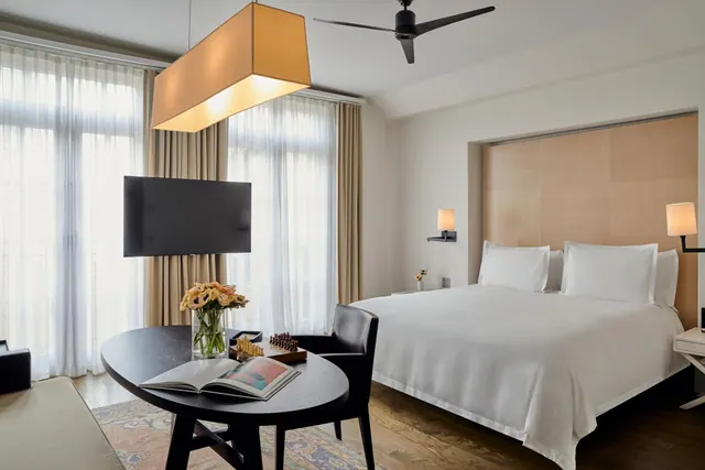 A bedroom with a king bed under white linens, a wall-mounted television, a round black table with flowers and an open book, and tall windows covered by sheer curtains.
