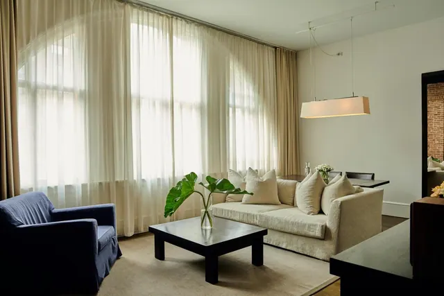 A living area with a beige sofa, a navy armchair, a black coffee table with a plant vase, and sheer curtains over tall windows.