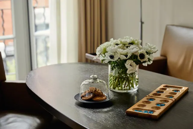 A round dark wood table holds a vase of white anemones, a glass cloche over cookies, and a wooden mancala board, with a leather armchair and a window beside it.