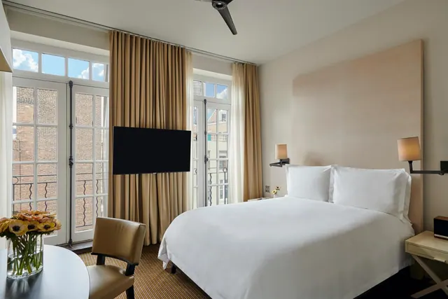 A bedroom with a made king bed, a wall-mounted television, and French doors looking out onto a brick building, with a round table holding a vase of flowers.