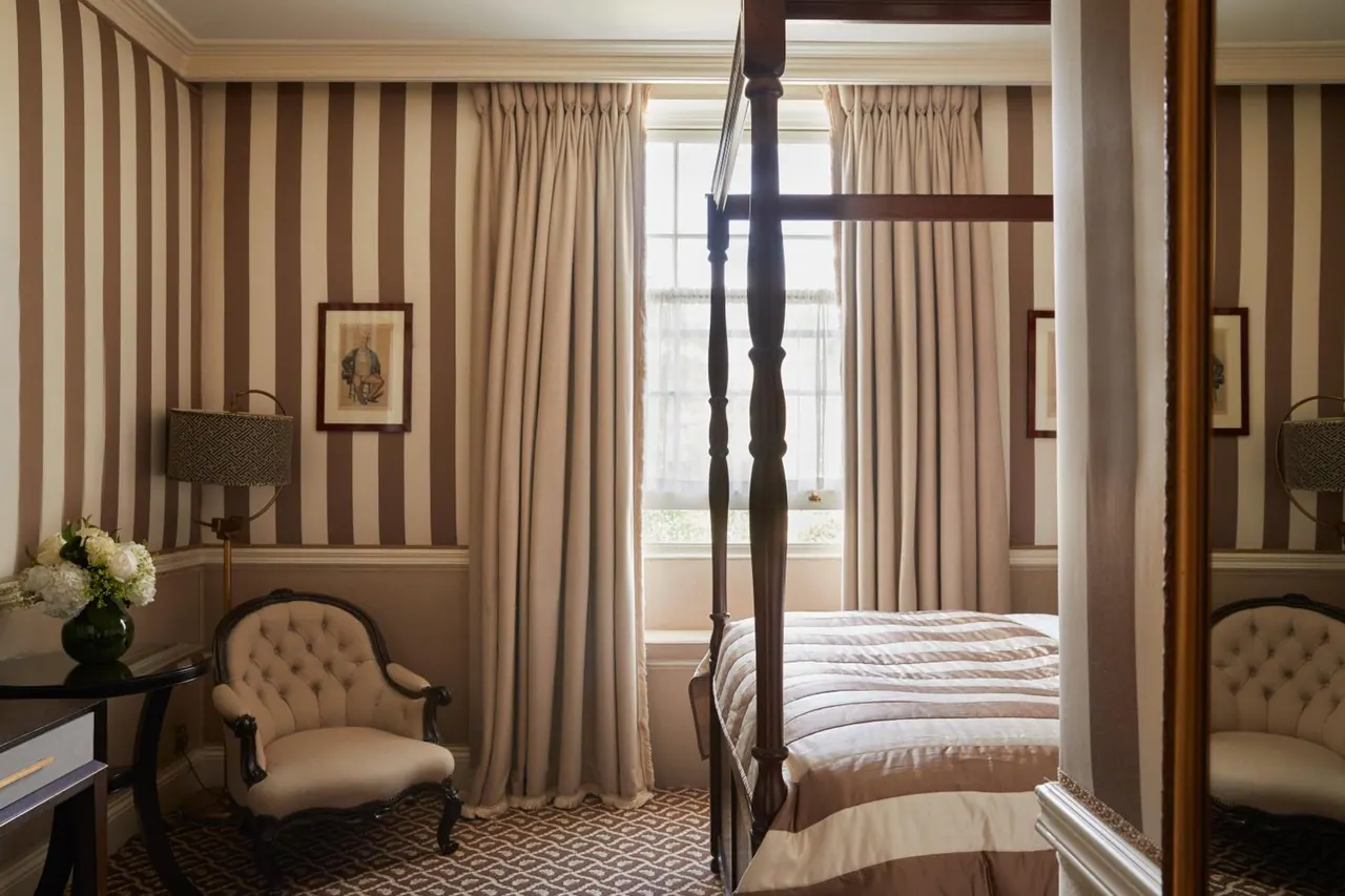 A bedroom with a four-poster bed, striped wallpaper, a tufted armchair and a window with long curtains.
