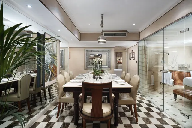 A long dining table is set for a meal in a room with a black and white checkerboard marble floor, and a glass-walled bathroom with a freestanding bath is visible on the right.