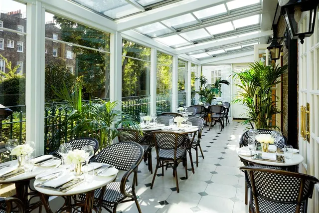 A glass-roofed dining room with round tables set with white flowers and glassware, with trees and brick buildings visible through the windows.