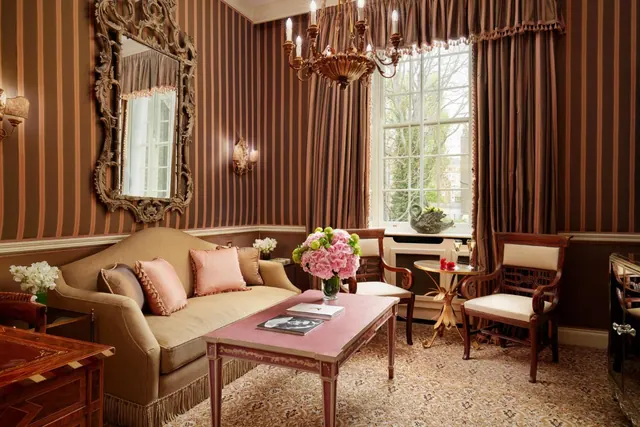 A living area with striped wallpaper, a tan sofa, a pink coffee table with a flower arrangement, a chandelier, and a window looking out onto bare trees.
