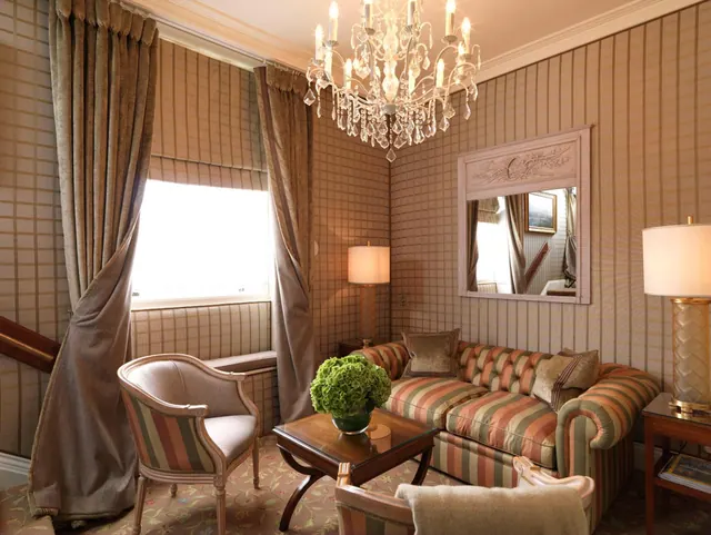 A living area with a striped sofa, armchairs, a crystal chandelier, table lamps and a framed mirror beside a curtained window.