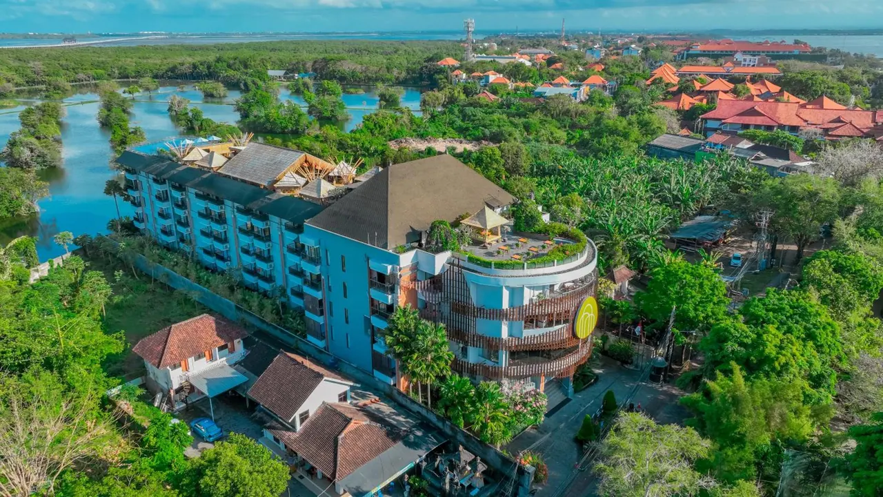 Aerial view of a blue multi-storey hotel with a curved wooden-balconied wing, surrounded by trees, a lagoon and red-tiled houses.