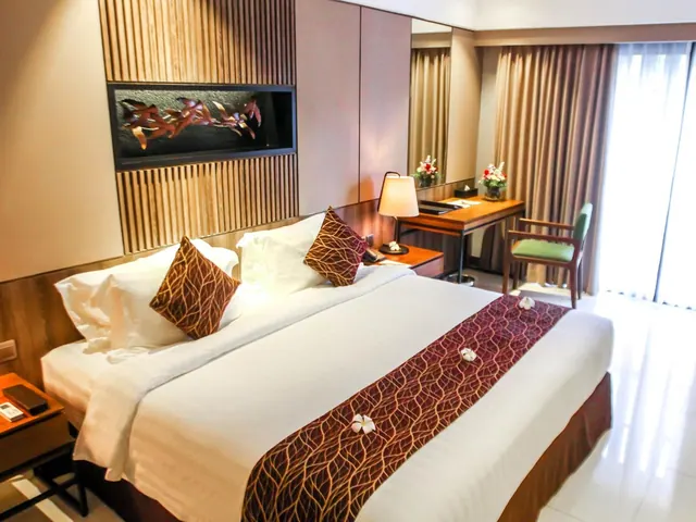 A king bed with white linens and a patterned runner sits beside a wooden desk, chair and lamp, with tall windows on the right.