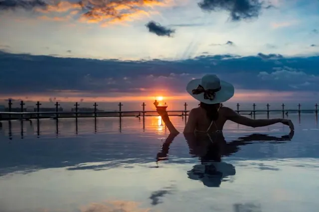A woman in a wide-brimmed hat holding a drink sits in an infinity pool facing the sea at sunset.