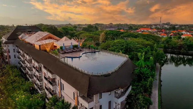 Aerial view of a multi-storey hotel with a thatched roof, a rooftop pool with loungers and parasols, a lake on the right, and a village at sunset.