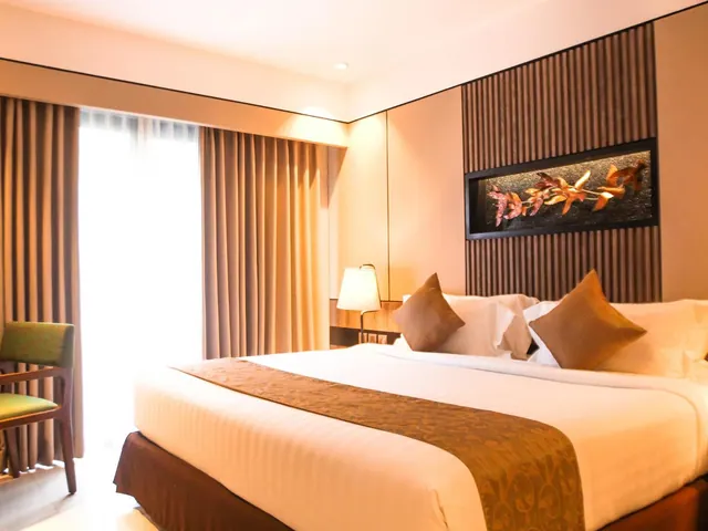 A hotel bedroom with a made king bed, a brown and gold bed runner, decorative pillows, a framed leaf artwork on a slatted wall, and curtains beside a bright window.