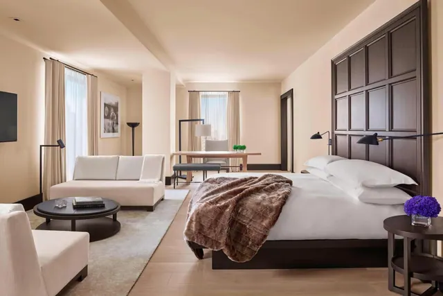 A bedroom with a king bed under a fur throw, a white sofa and armchair beside a round coffee table, and a desk near the tall windows.