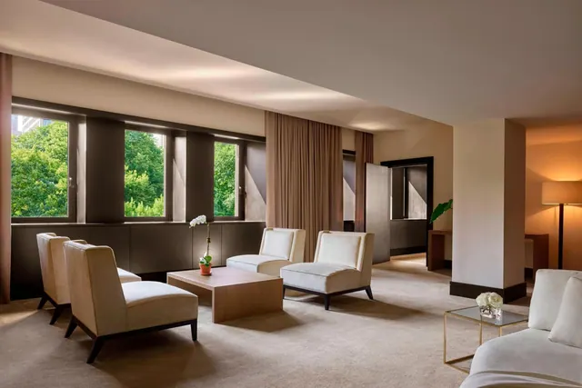 A suite living area with cream armchairs, a wooden coffee table, a floor lamp and windows looking out onto green trees.
