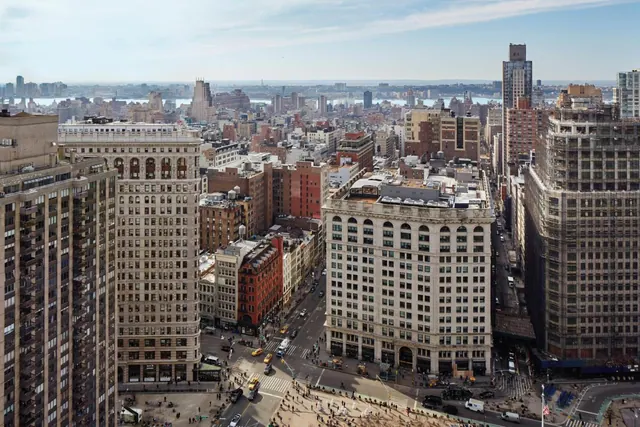 Aerial view looking down on dense city blocks, streets and traffic, with a body of water visible on the horizon under a pale sky.