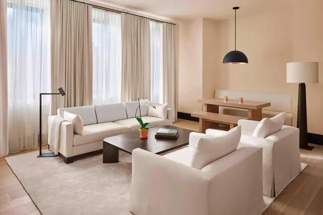 A furnished living room with a white sofa, two white armchairs, a dark coffee table, a dining table with bench, and sheer-curtained windows.