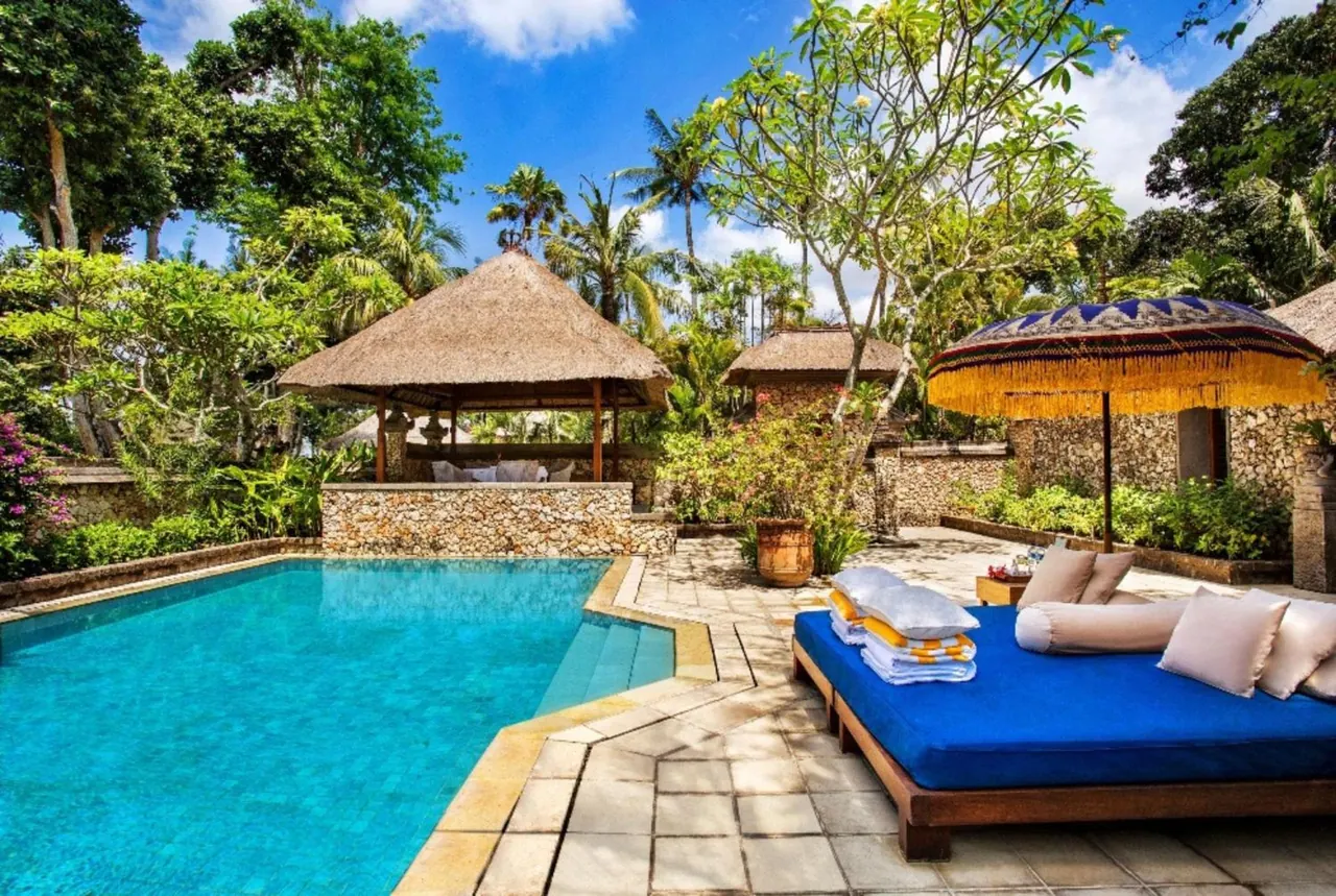 A swimming pool with a thatched gazebo, a yellow-fringed umbrella and a blue daybed with cushions beside it, surrounded by tropical trees.