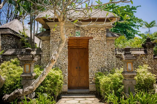 A stone-walled entrance with a thatched roof, carved wooden double doors, stone lanterns and a frangipani tree in front.