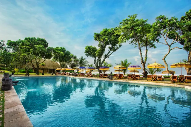 A large blue swimming pool with stone spouts on the left, lined with sun loungers and purple and gold umbrellas, with trees and the sea beyond.