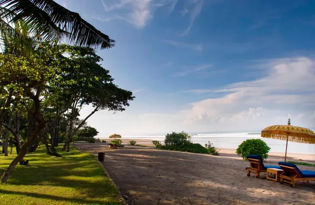 Blue-cushioned sun loungers and thatched umbrellas stand on a sandy beach beside a grassy lawn, with the sea and waves visible behind them.