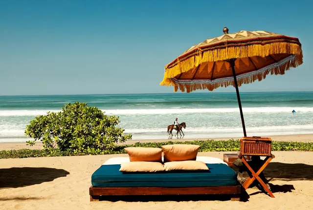 A daybed with cushions, a fringed Balinese parasol and a wicker side table stand on sand facing the sea, with a horse and rider on the beach.