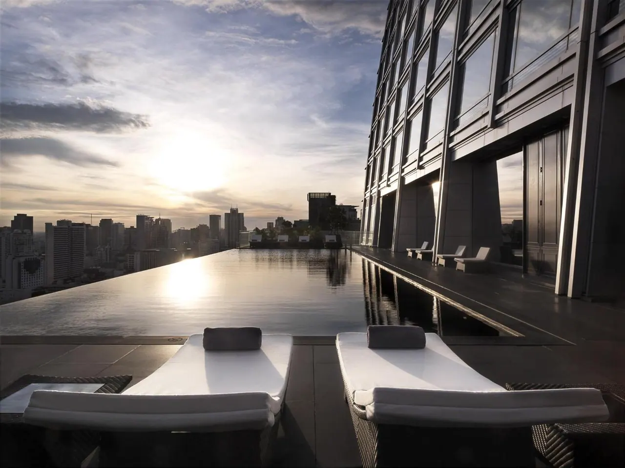 An infinity-edge rooftop pool with white sun loungers overlooks a city skyline at sunset.