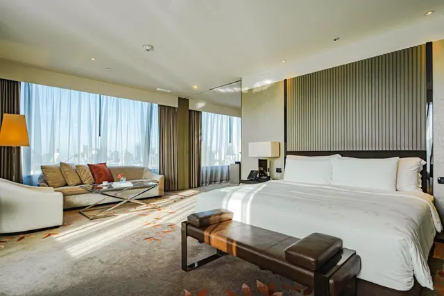 A bedroom with a king bed, a leather bench at its foot, and a sofa area by floor-to-ceiling windows looking out over the city.