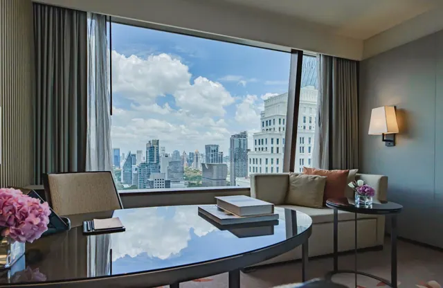 A hotel room living area with a grey sofa, a round glass table and a large window looking over a city skyline under a partly cloudy sky.