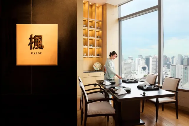 A staff member in a kimono sets a table beside floor-to-ceiling windows with a city skyline view, next to a wall sign reading KAEDE.