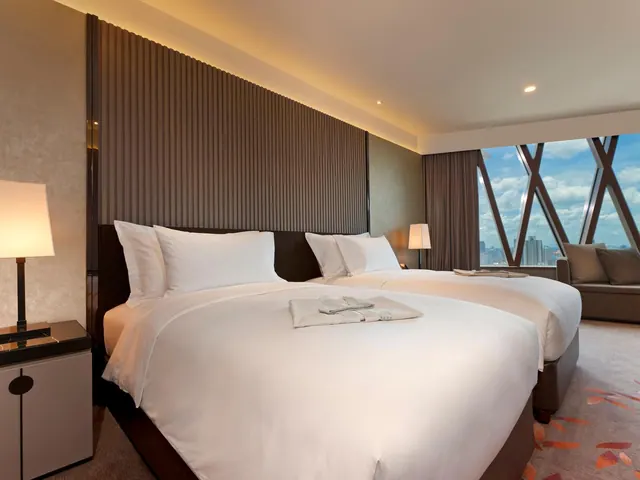 A bedroom with two beds dressed in white linen, a sofa at the right, and a large window looking out over city buildings under a cloudy sky.