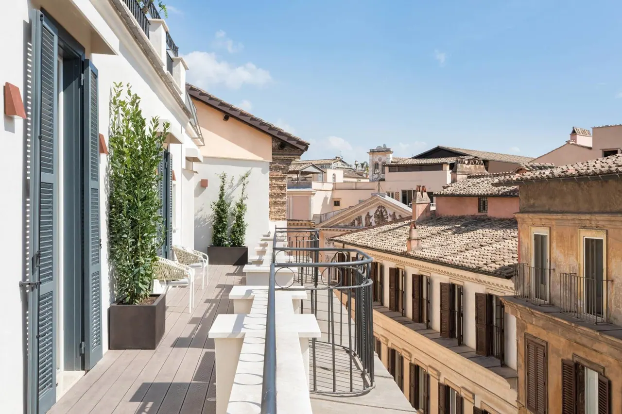 A sunlit wooden-decked rooftop terrace with white chairs, potted shrubs, a curved metal railing and grey shutters, overlooking tiled rooftops and a bell tower under a blue sky.