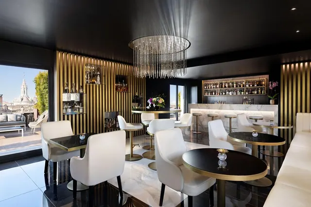 A dark-ceilinged lounge with gold slatted walls, a crystal chandelier, white leather chairs and round tables, a bar along the back wall, and a terrace with a church view visible through the left window.