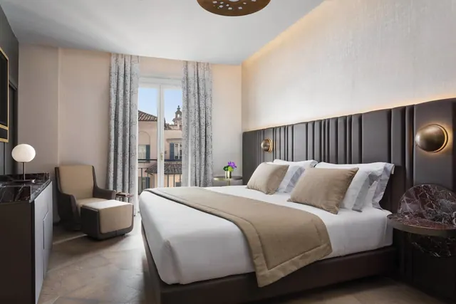 A bedroom with a king bed, an armchair and ottoman, and glass doors opening onto a balcony with a view of rooftops and a bell tower.
