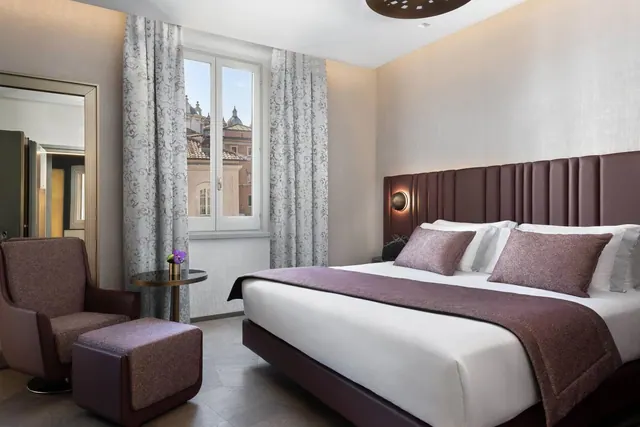 A staged bedroom with a large bed, an upholstered armchair and ottoman, and a window showing rooftops and domes outside.