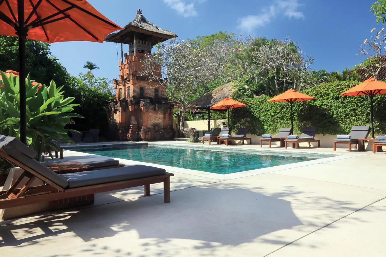 A rectangular swimming pool with sun loungers and orange parasols on the pool deck, with a brick Balinese tower and trees behind.