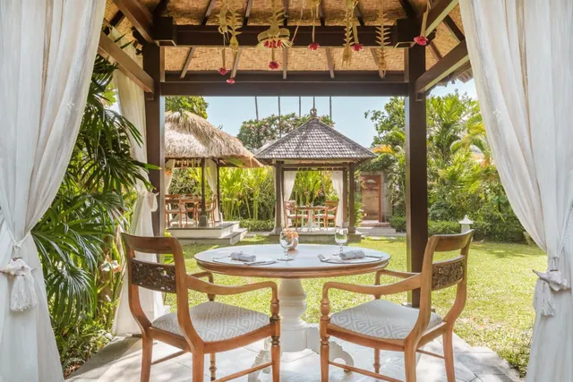 An open-sided thatched pavilion with a round white table and two wooden chairs looks out onto a lawn and other garden pavilions.