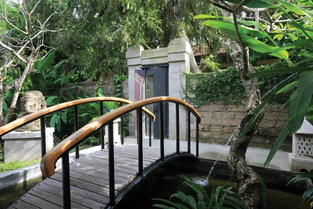A curved wooden footbridge with black metal railings crosses a garden pond, with a stone statue and a carved stone gateway behind it.
