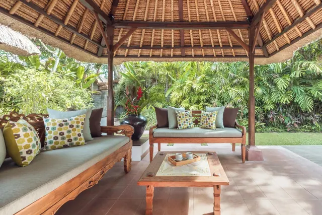 A covered thatched pavilion with two wooden daybeds, cushions, a low coffee table with a tray and cups, and a lush garden behind.