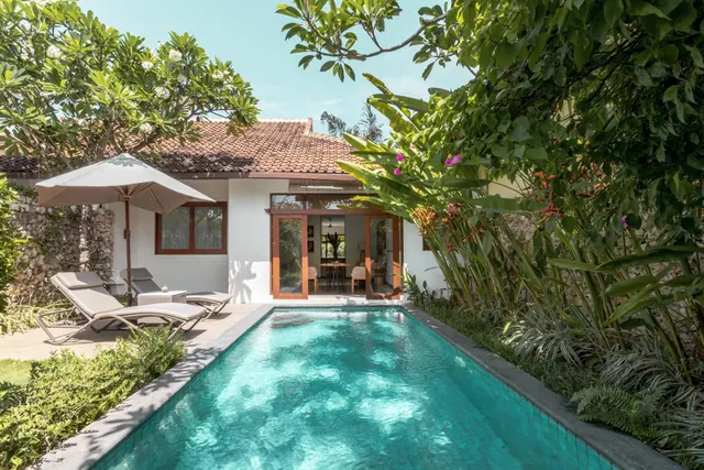 A narrow turquoise swimming pool runs beside a white bungalow with a terracotta tiled roof, with two sun loungers and a parasol on the deck and lush tropical planting around it.