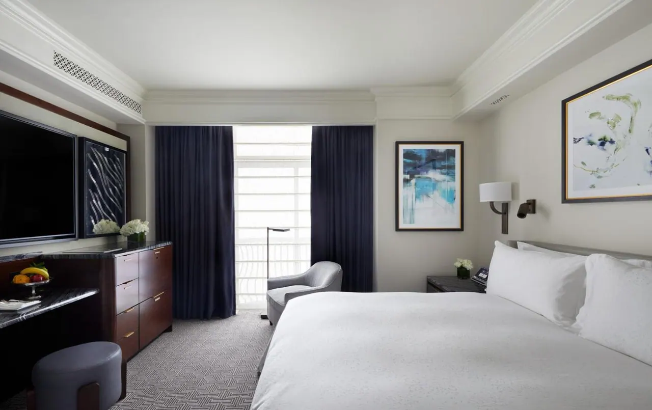 A bedroom with a made king bed, a wall-mounted television, a wooden dresser, and a grey armchair beside a window with navy curtains.